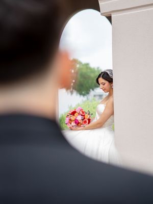 Creative wedding photo of bride holding pink bouquet viewed through arched columns with groom in foreground.