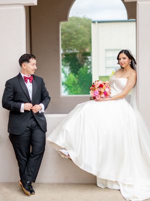 Bride in white ball gown sitting on ledge with bouquet while groom in black tuxedo stands nearby.