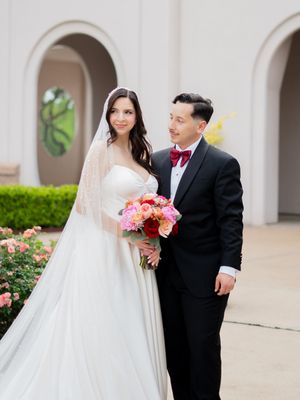 Wedding couple posing outdoors near rose garden, bride in pearl veil and groom in black tuxedo.