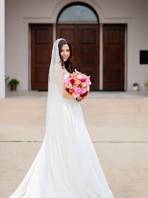 Stunning bride in white gown with long flowing veil holding colorful bouquet before church wooden doors.