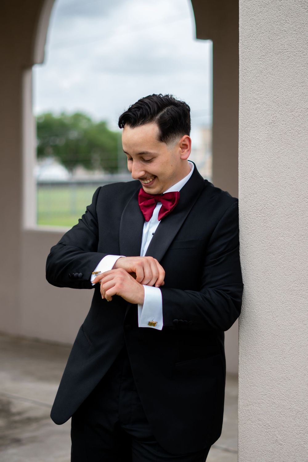 Dapper groom in black tuxedo with burgundy bow tie smiles while checking his cufflinks before the wedding ceremony.
