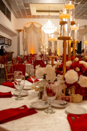 Elegant wedding reception hall with red and white floral centerpieces, gold candelabras, and chandelier.