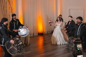 Bride and groom enter wedding reception accompanied by dhol drummers in traditional attire on a wooden dance floor.