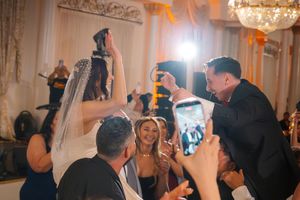 Bride and groom celebrate with guests on the dance floor during a lively wedding reception under chandelier lighting.