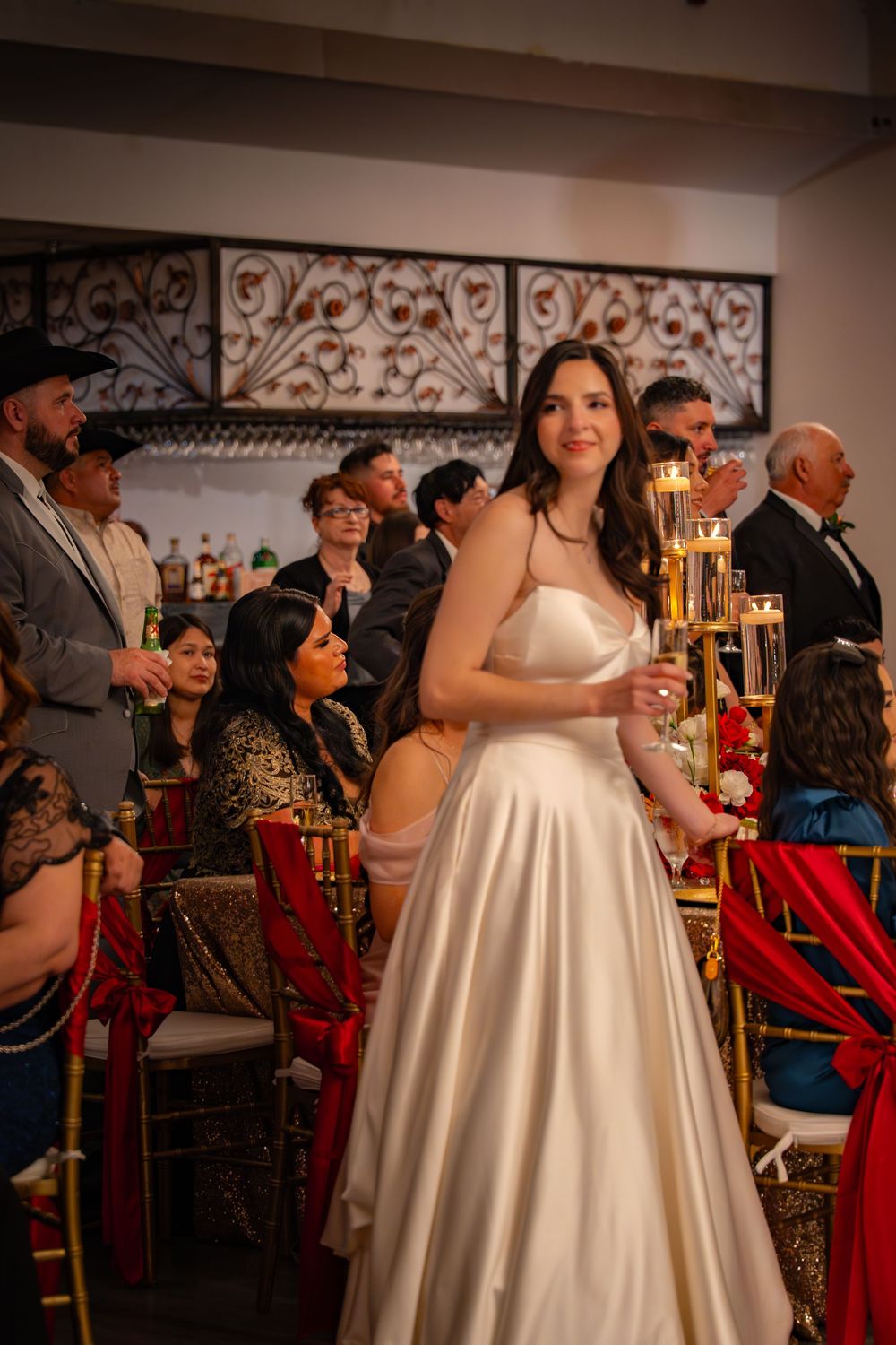 Elegant bride in a satin gown holds a champagne glass while standing among seated wedding reception guests.