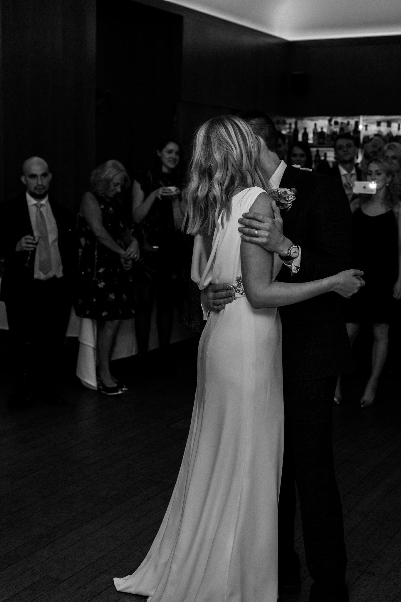 Black and white photo of a bride and groom sharing their first dance at a wedding reception with guests watching.
