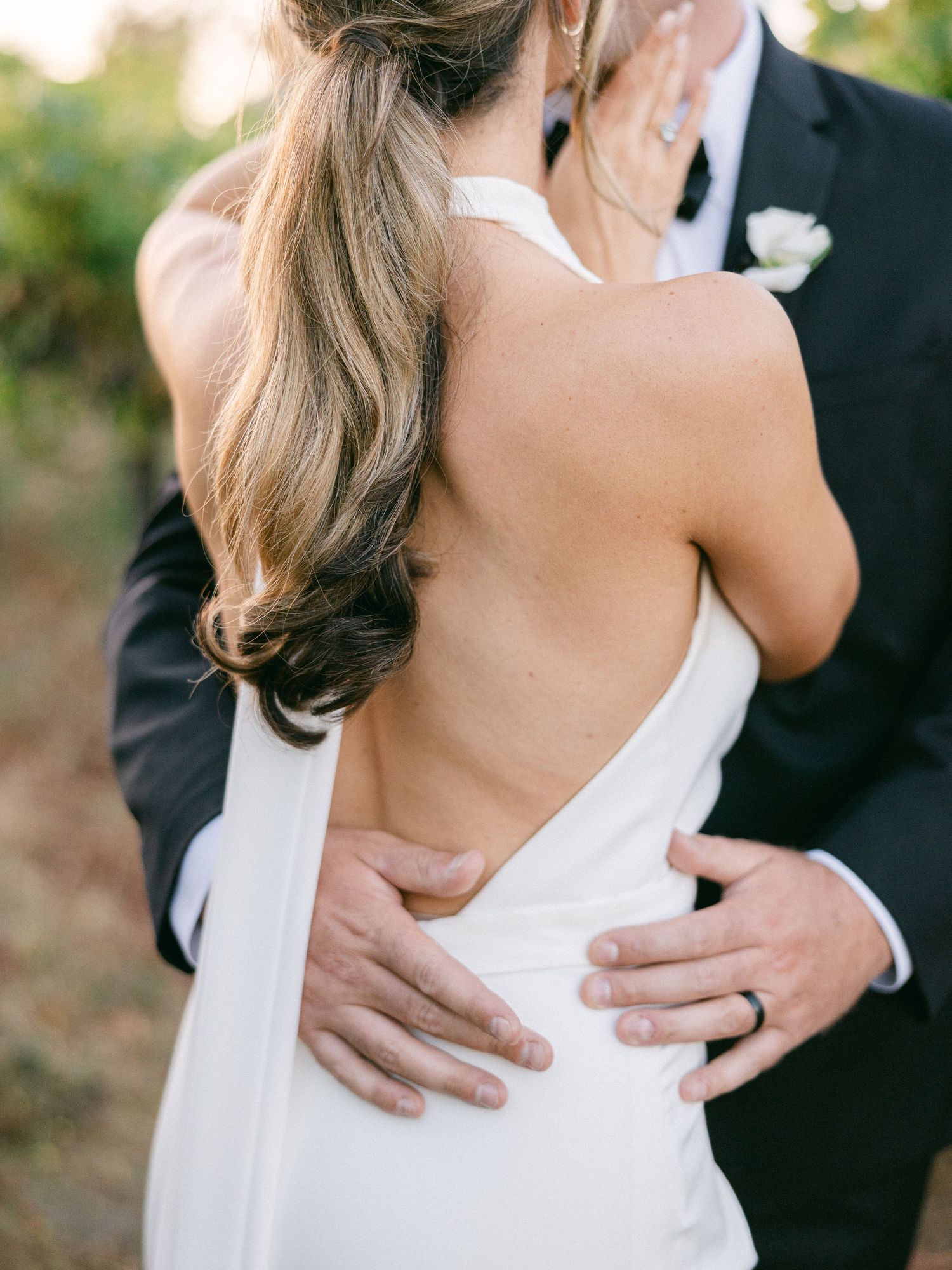 Bride in backless white halter gown embraced by groom in black suit at outdoor wedding ceremony.