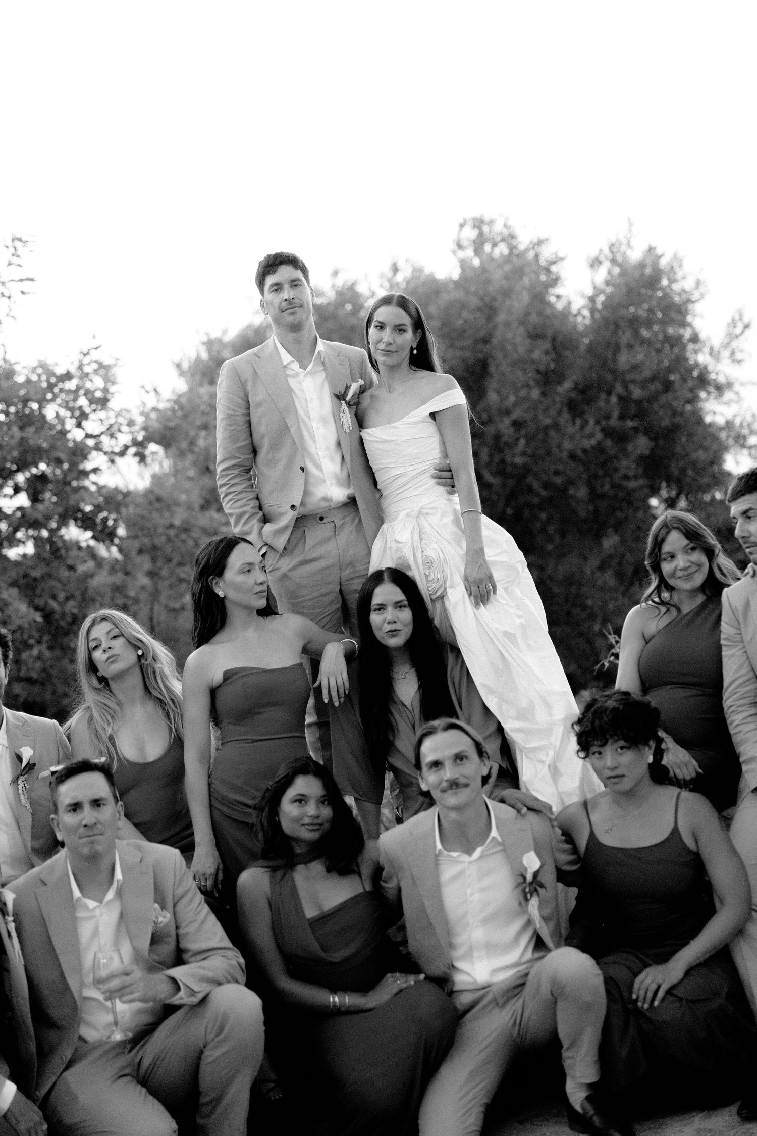 Black and white wedding group photo with bride and groom elevated above smiling guests outdoors.