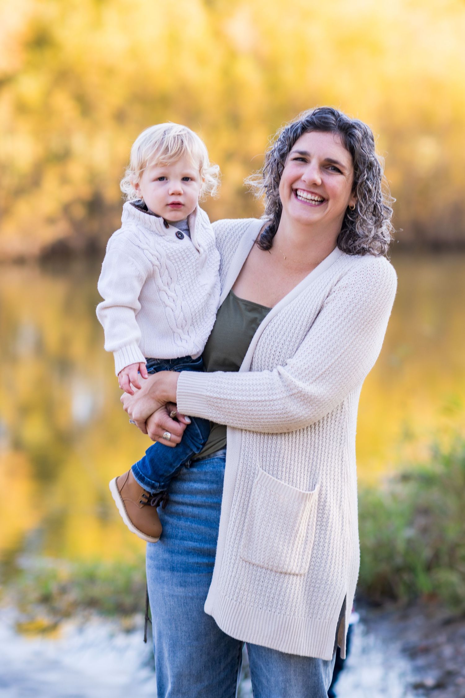A smiling woman with curly hair holds a toddler in a white sweater outdoors near a pond with autumn foliage.