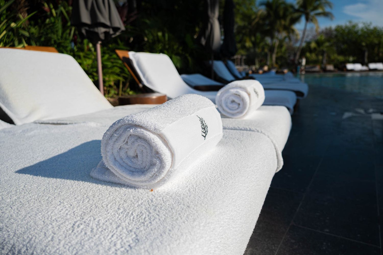 Rolled white towels on a luxury poolside lounge chair at a tropical resort with palm trees in the background.