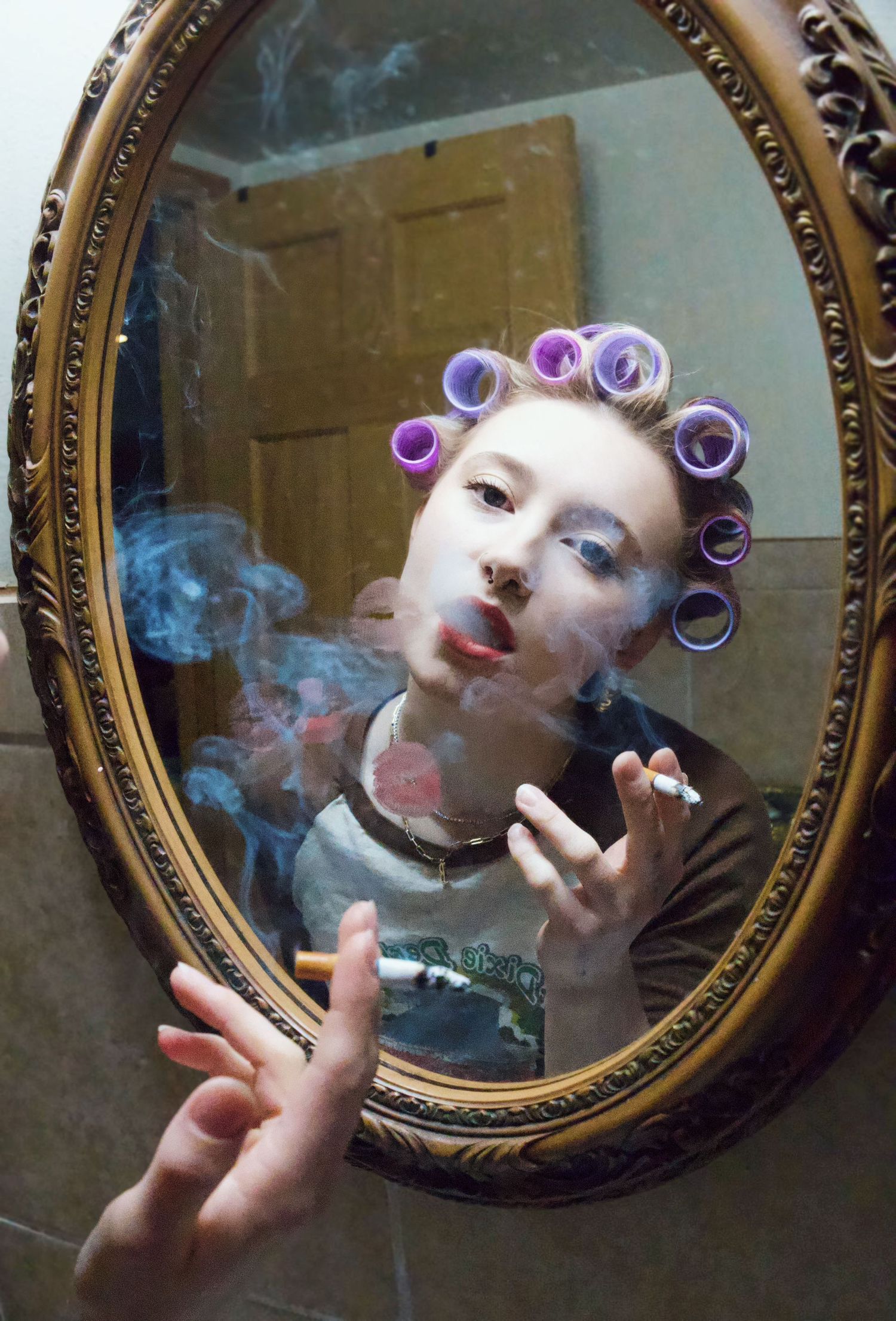 Person with purple hair rollers smoking a cigarette, reflected in an ornate oval gold mirror.