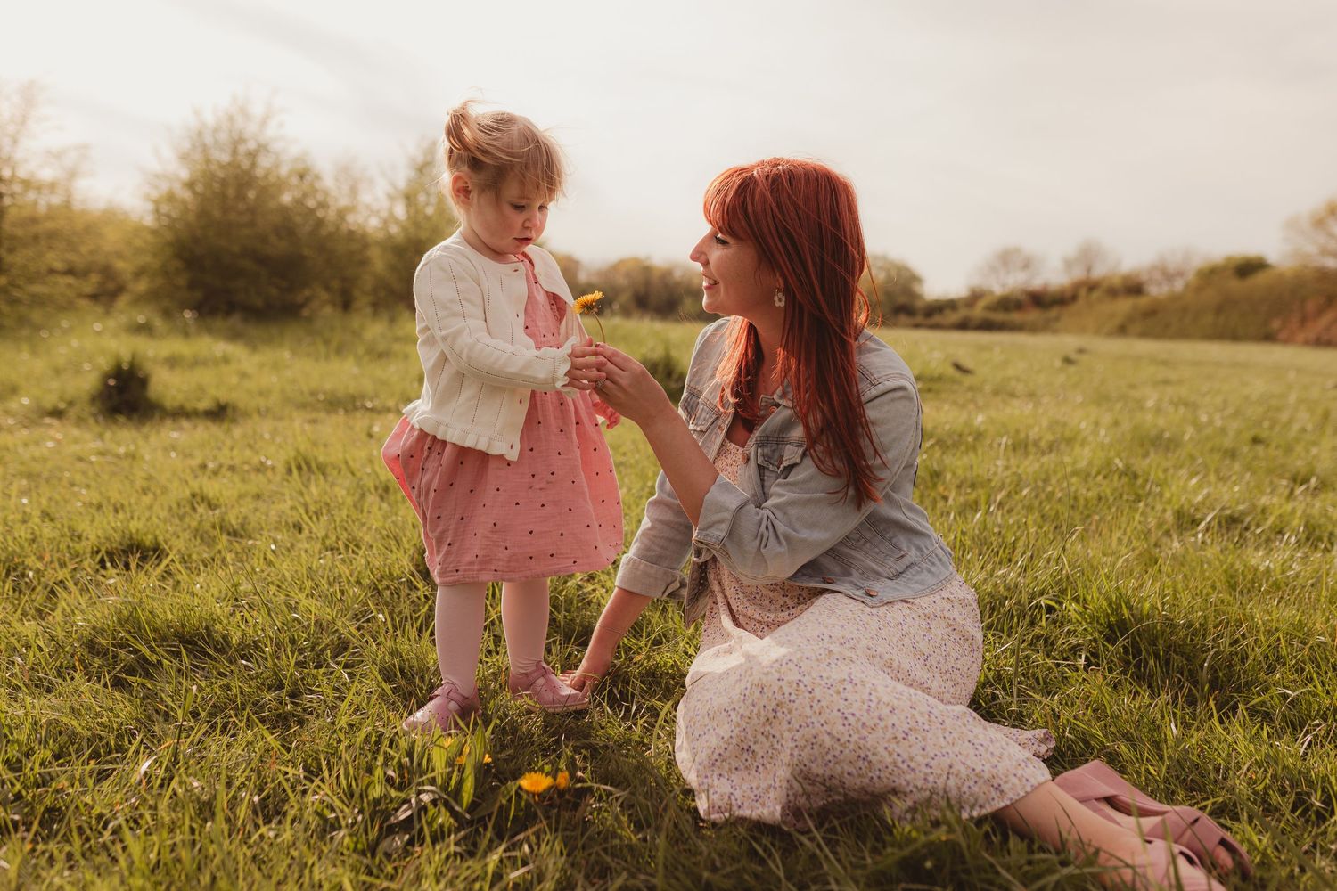 A red-haired mother kneels on green grass, sharing a yellow flower with her toddler daughter in a sunny field.