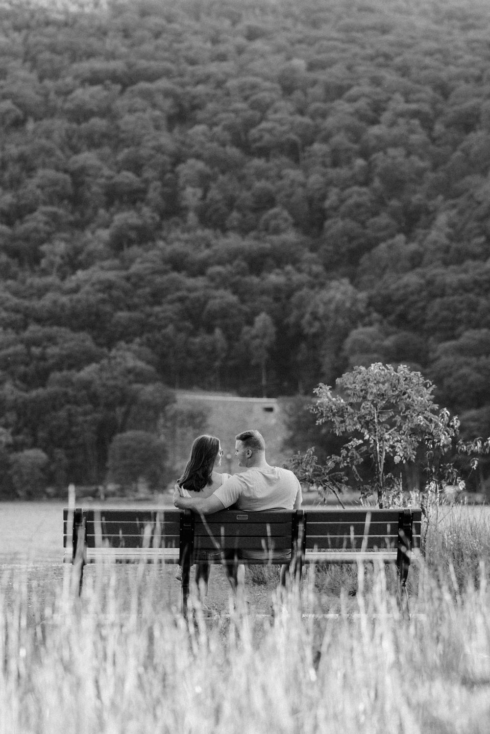Black and white photo of a couple sitting on a bench, embracing while overlooking a lush forested hillside.