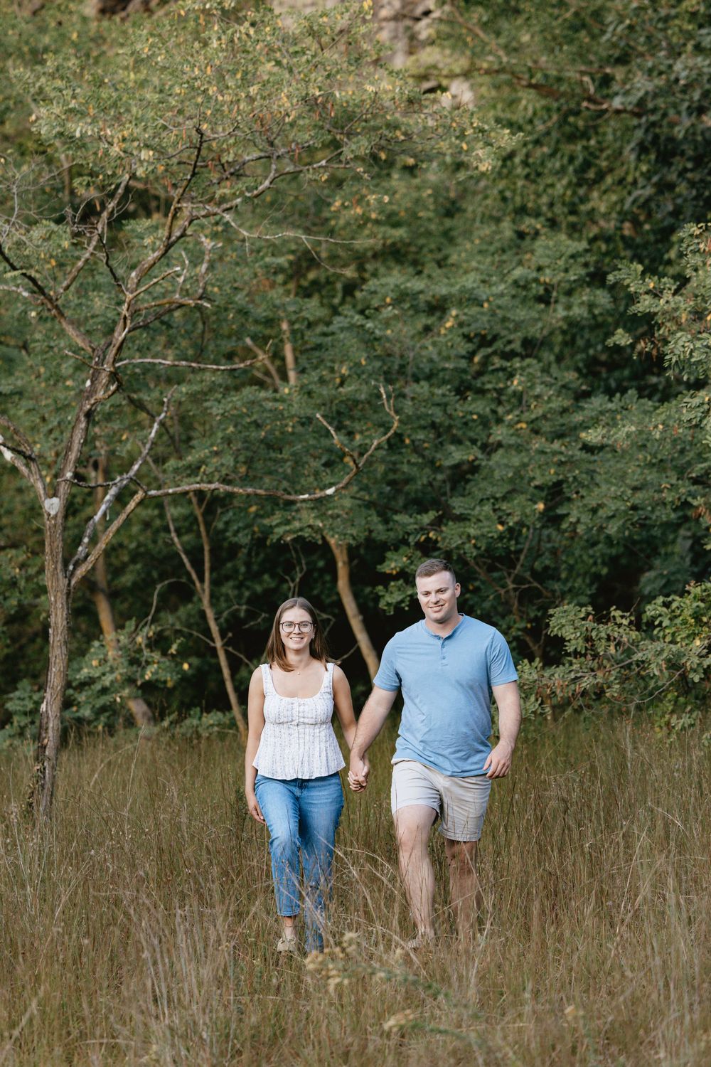 A couple holds hands while walking through tall grass with lush green trees in the background.