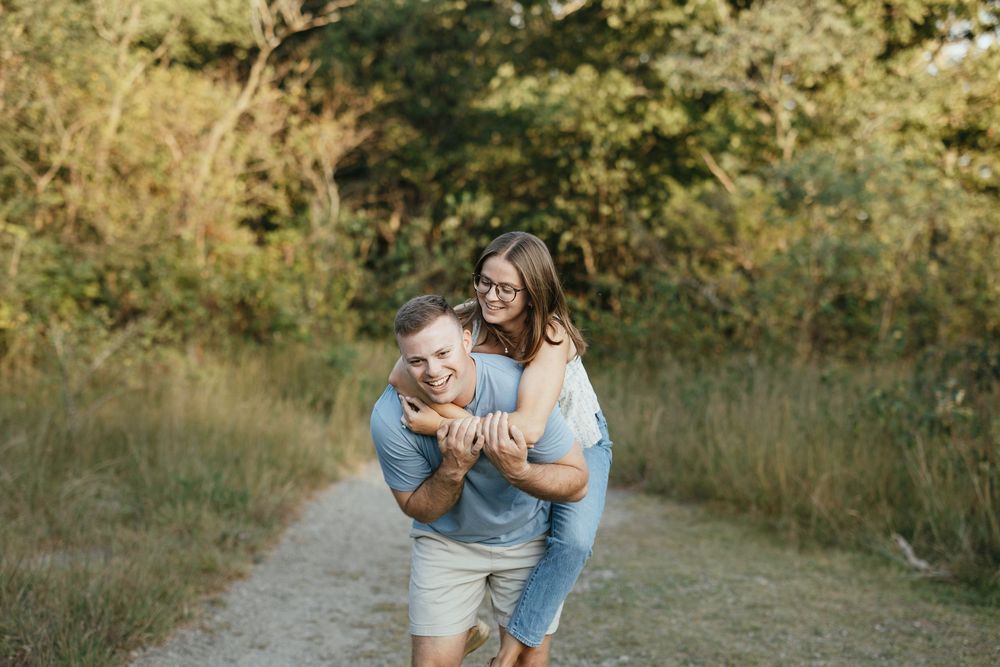 Couple laughing outdoors on a dirt path, man giving woman a piggyback ride surrounded by lush greenery.