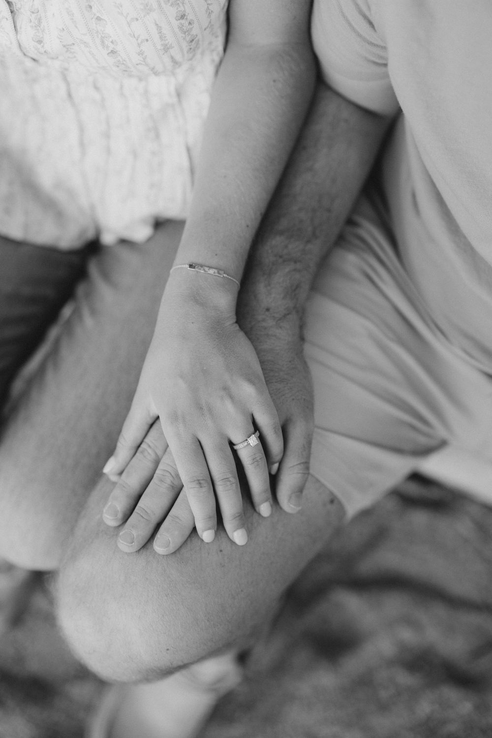 Black and white photo of a couple's hands resting together, showing an engagement ring and bracelet on a knee.