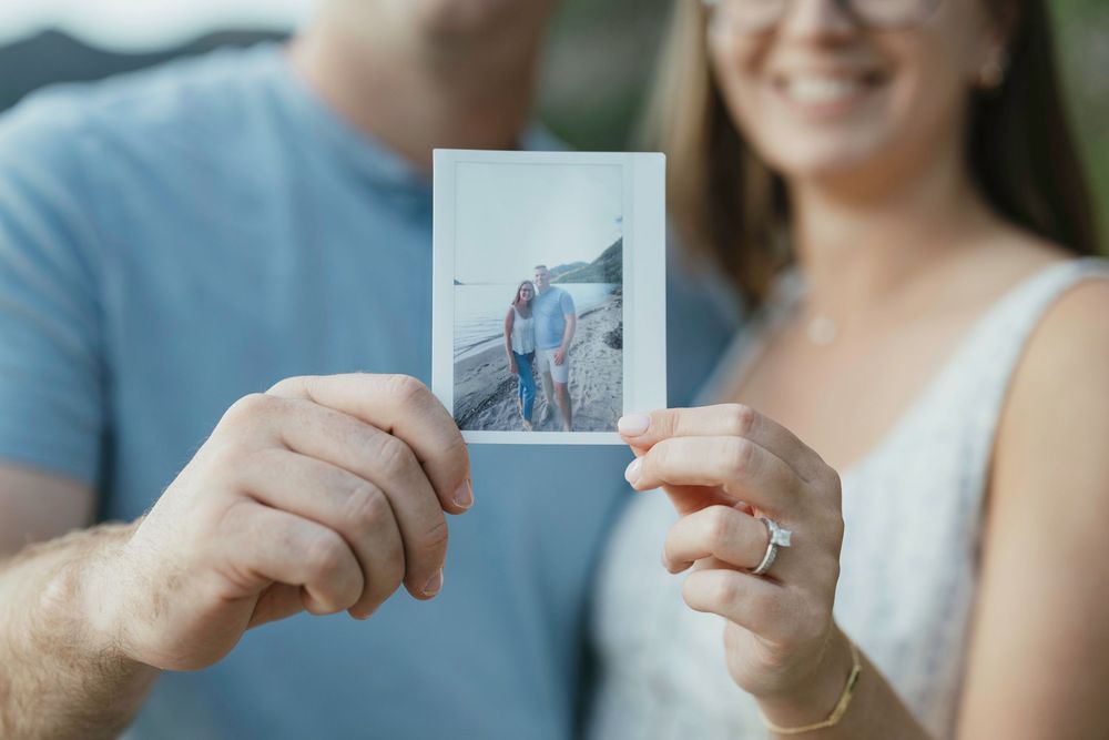 Couple holding a Polaroid photo of themselves standing on a beach, smiling and wearing casual outfits.
