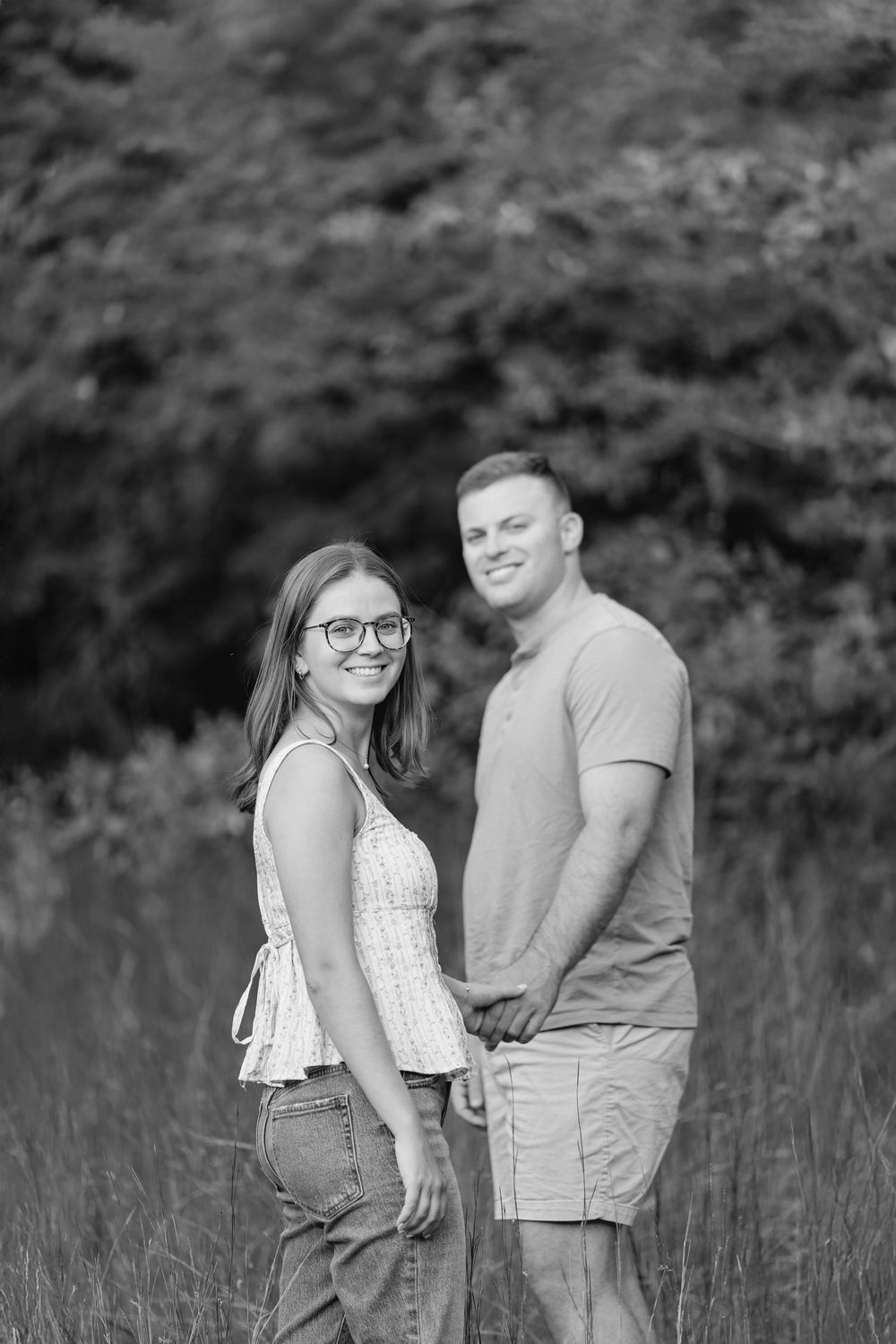 Black and white photo of a smiling couple holding hands in a field with trees in the background.