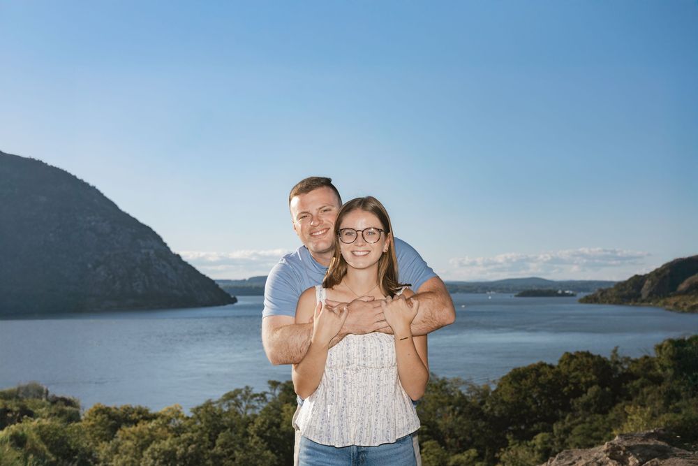 Couple embracing outdoors with scenic lake and green hills under a clear blue sky.