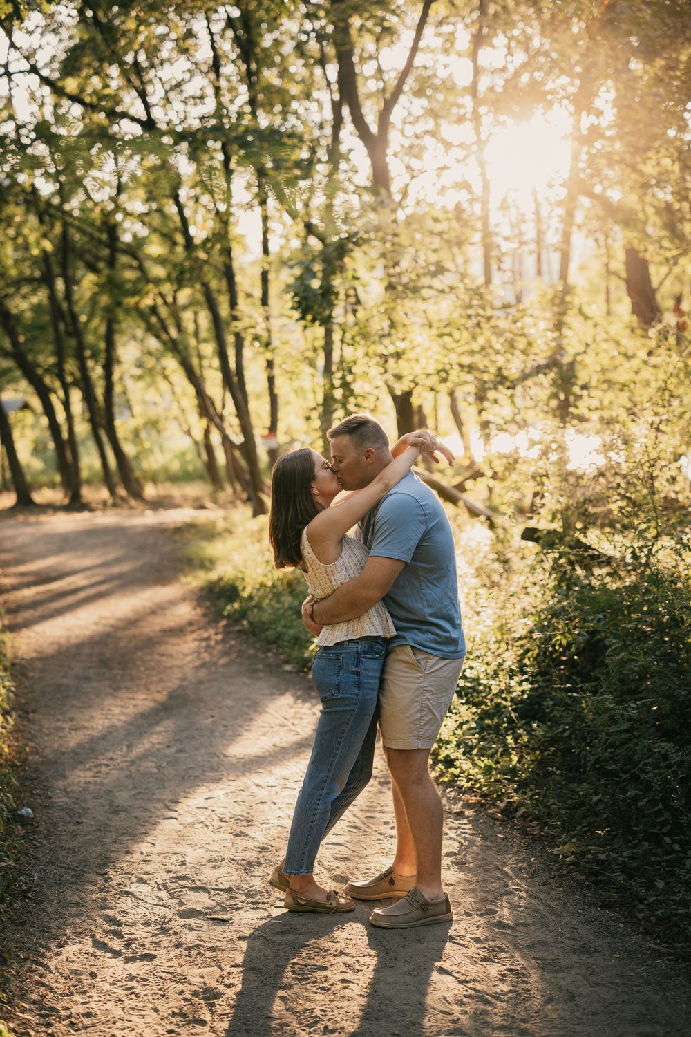 Couple embracing and kissing on a sunlit forest path surrounded by lush green trees during golden hour.