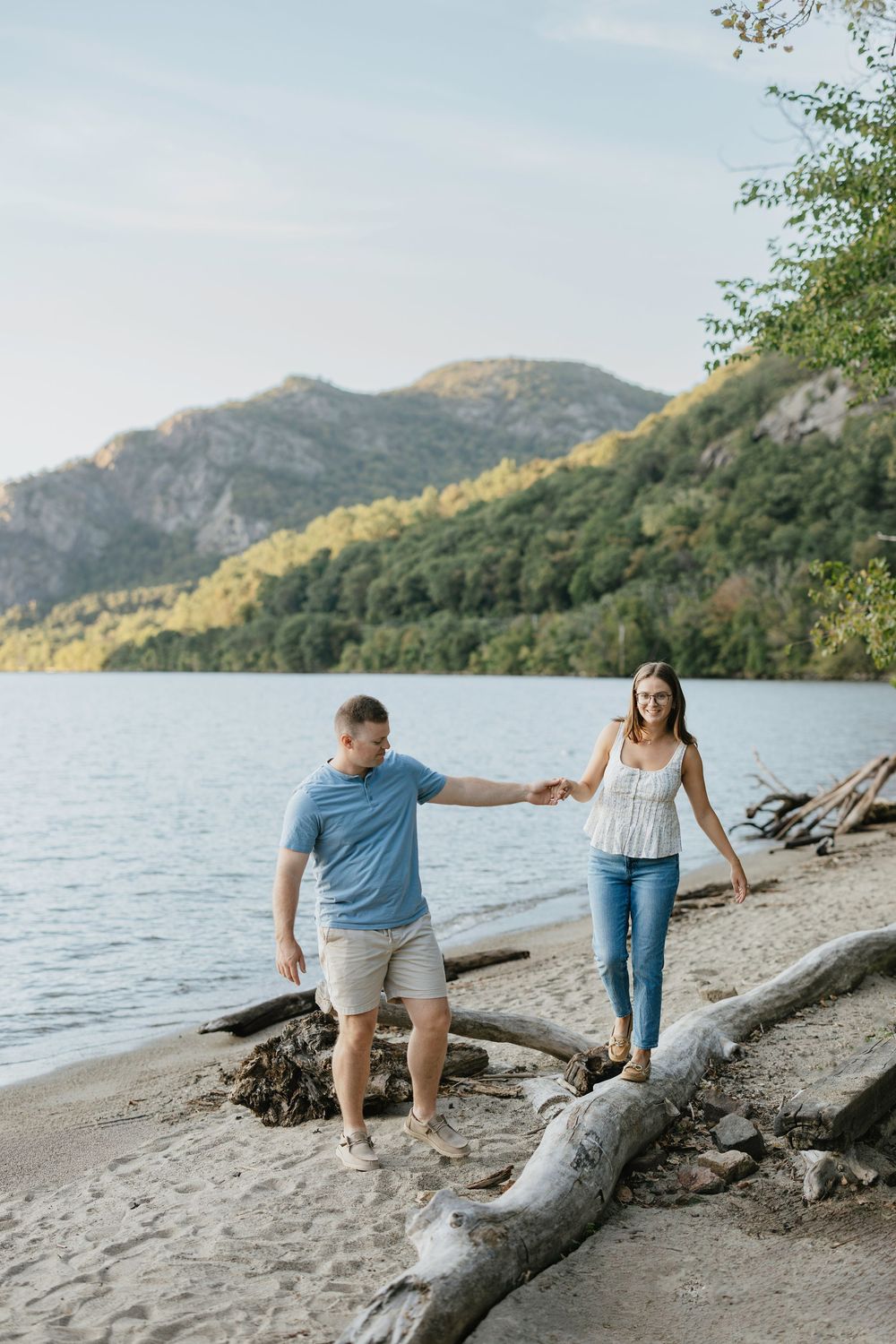 Couple holding hands walking along a sandy lakeside beach with mountains and trees in the background.