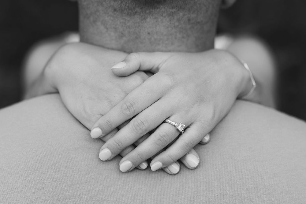 Black and white photo of two hands clasped together, one wearing a diamond engagement ring.