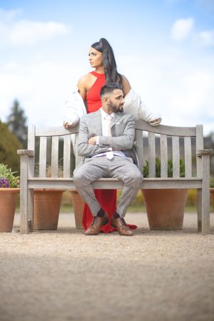 Elegant couple posing on a wooden bench outdoors, woman in red dress standing behind seated man in grey suit.