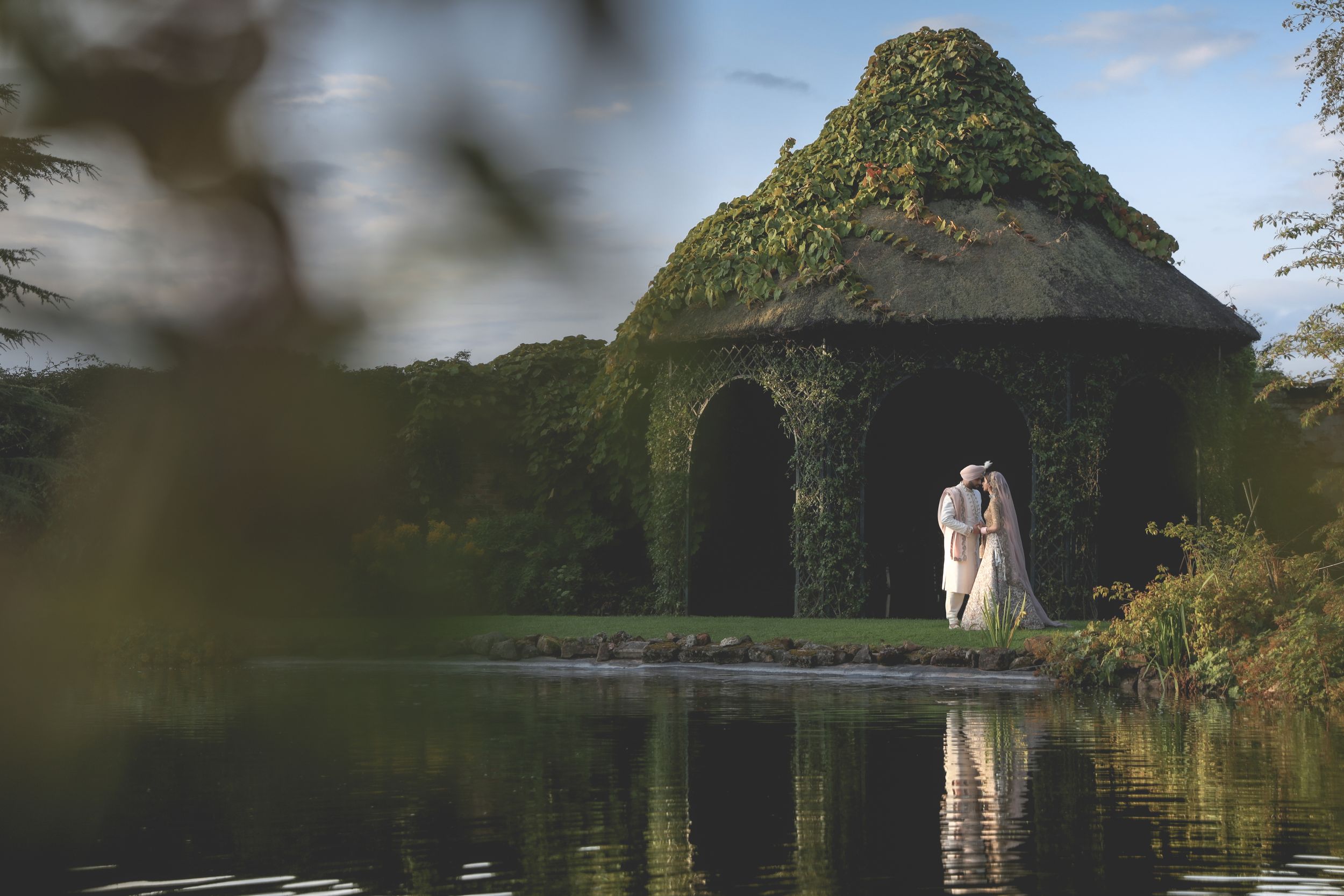 Couple embracing by ivy-covered chapel reflected in still pond at romantic garden wedding venue.