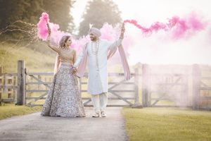 Indian couple in traditional attire holding pink smoke bombs on a garden path, celebrating their wedding day.