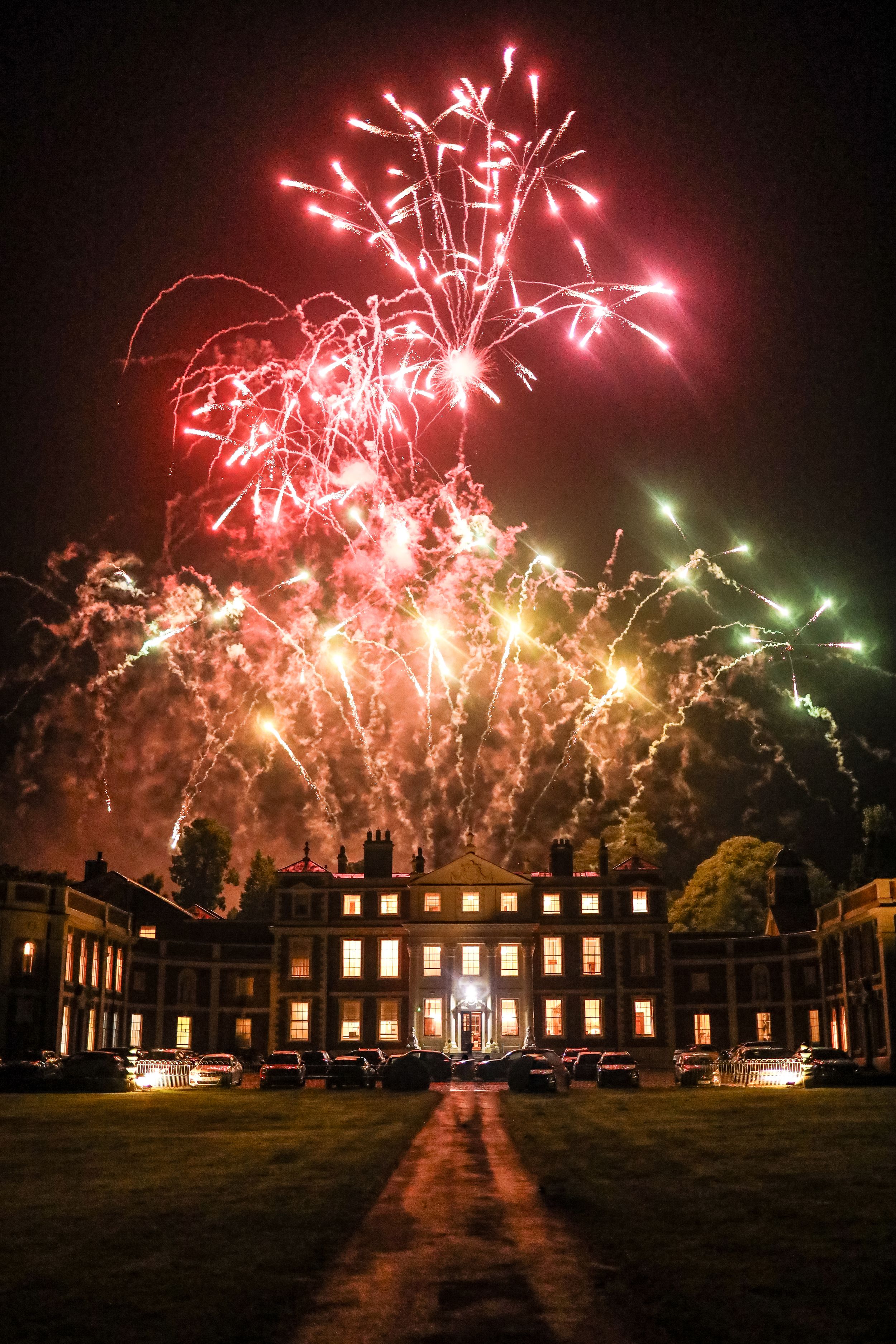 Spectacular fireworks display over a grand historic mansion at night, illuminating the sky in red and green bursts.