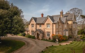 Historic stone manor house with Gothic architecture, gravel driveway, and lush gardens under a blue sky.
