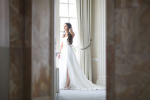 Elegant bride in a flowing white wedding gown standing by a large window in a luxurious marble interior.