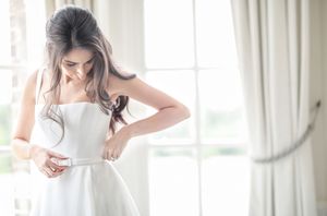 Bride in strapless white wedding gown adjusting her belt by a bright window with sheer curtains.