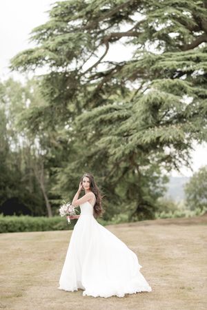 Bride in flowing white gown holding bouquet stands before large cedar tree in lush garden setting.