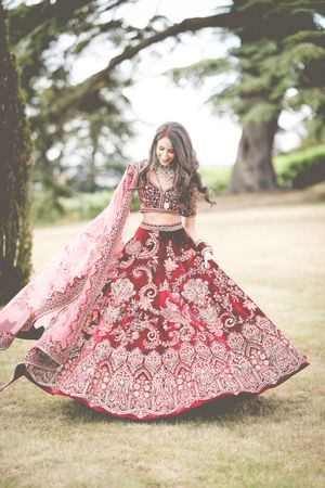 Woman twirling in ornate red and pink bridal lehenga with intricate embroidery outdoors under trees.