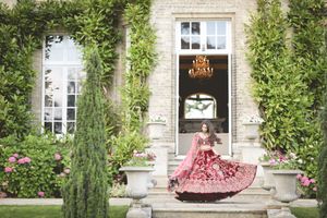 Bride in stunning red bridal lehenga twirling on steps of elegant stone mansion surrounded by lush greenery.