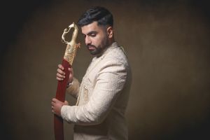 Man in ornate white sherwani holding a decorative gold and red sword, posing against a dark brown background.