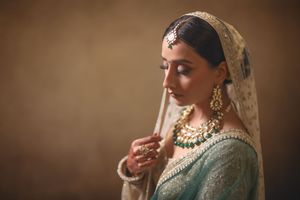 Indian bride in teal lehenga with gold jewelry and dupatta, posing gracefully in soft studio lighting.