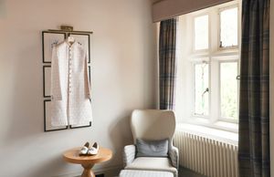 Elegant hotel room with a white dress on wall display, plaid curtains, and a cozy armchair by a window.
