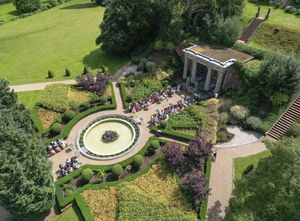 Aerial view of a formal English garden with a circular fountain, manicured hedges, and guests gathered near a classical stone pergola.