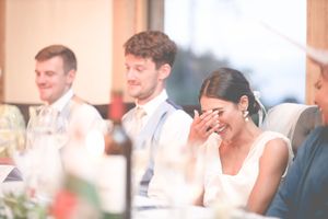 Bride laughs emotionally during wedding speeches at the reception dinner table with guests in background.