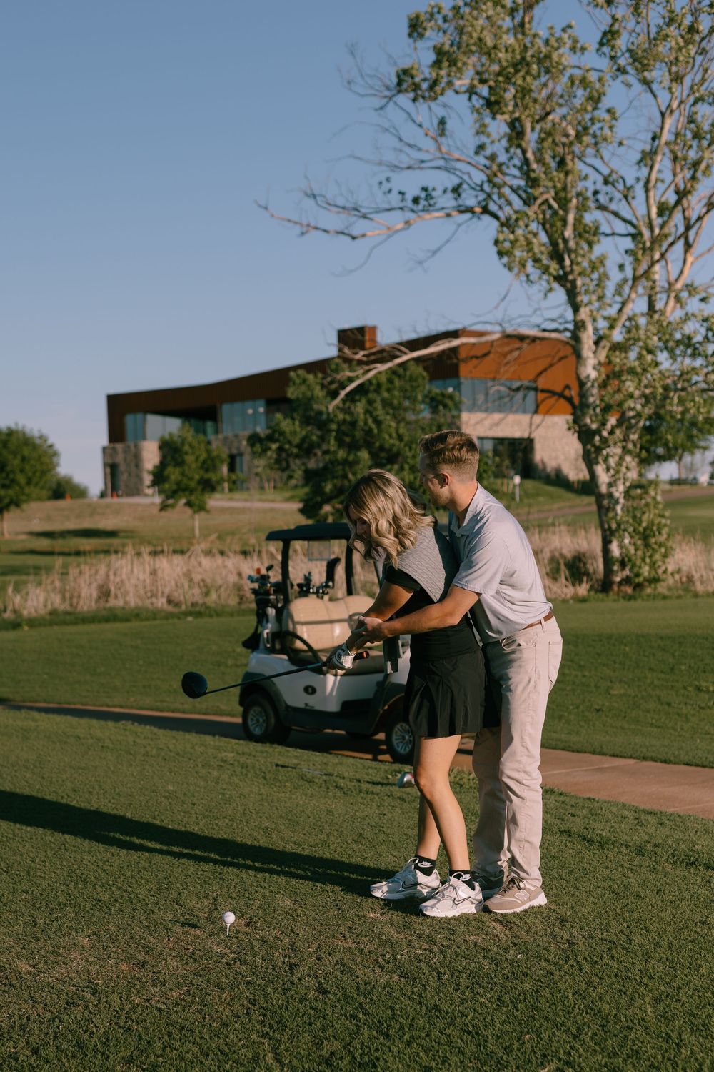 Couple sharing a golf lesson on a sunny course near a golf cart, with a modern building and trees in the background.