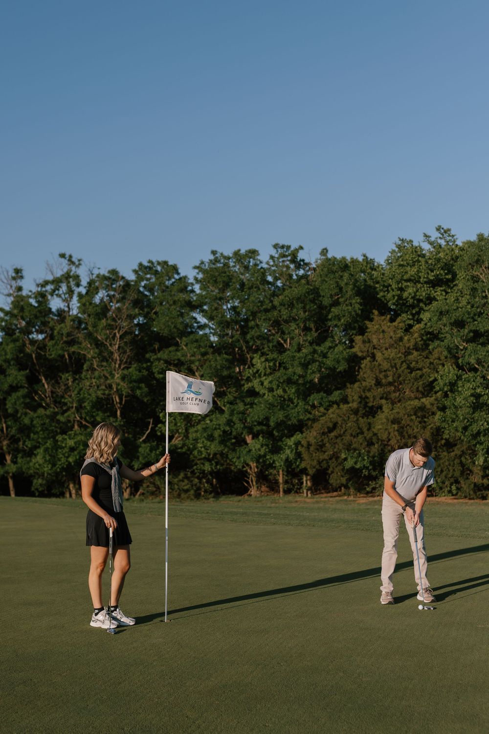 Two golfers on a sunny green, woman holding flag while man putts near a tree-lined course.
