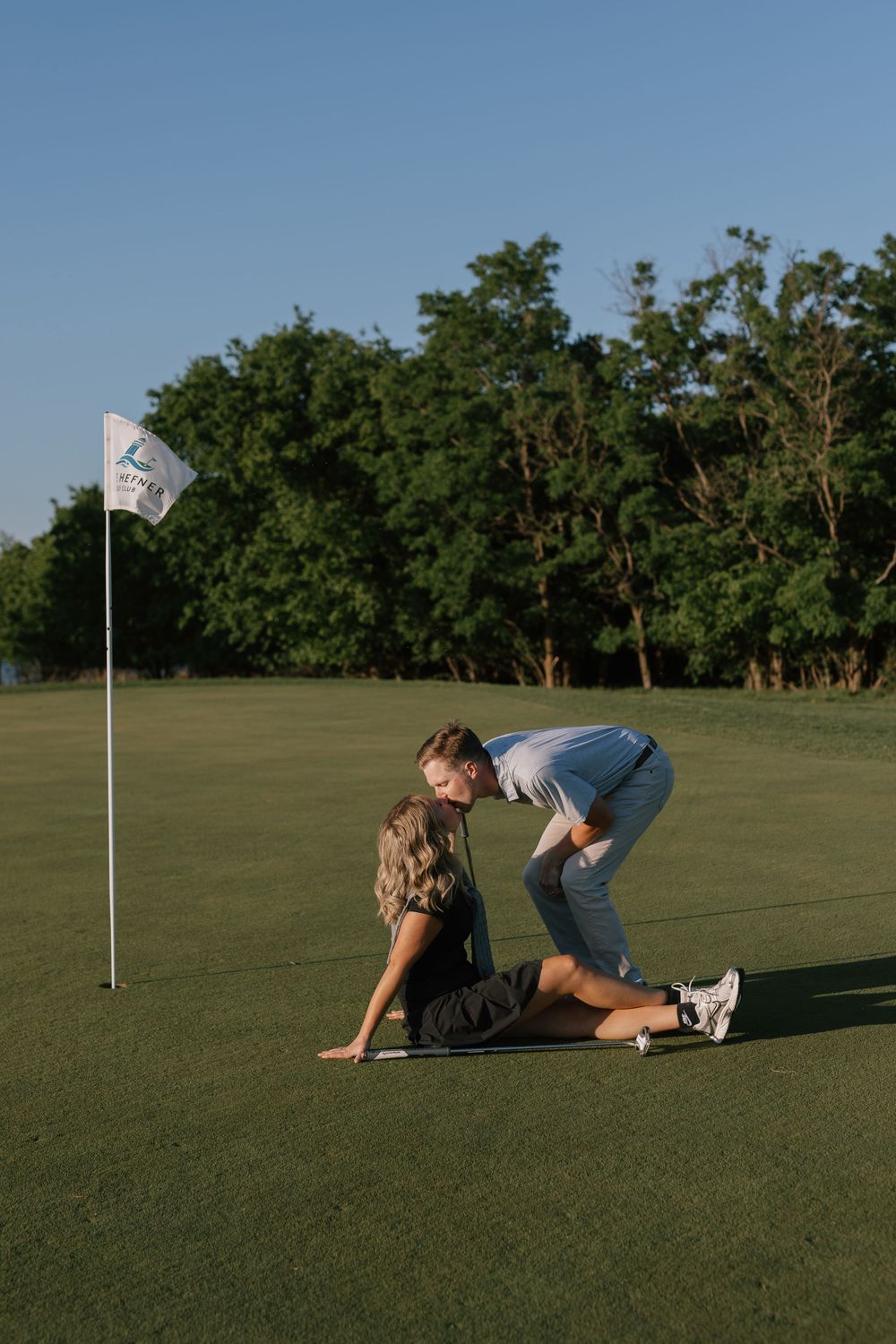 Couple sharing a romantic moment on a golf course green near a flag, surrounded by lush trees at sunset.