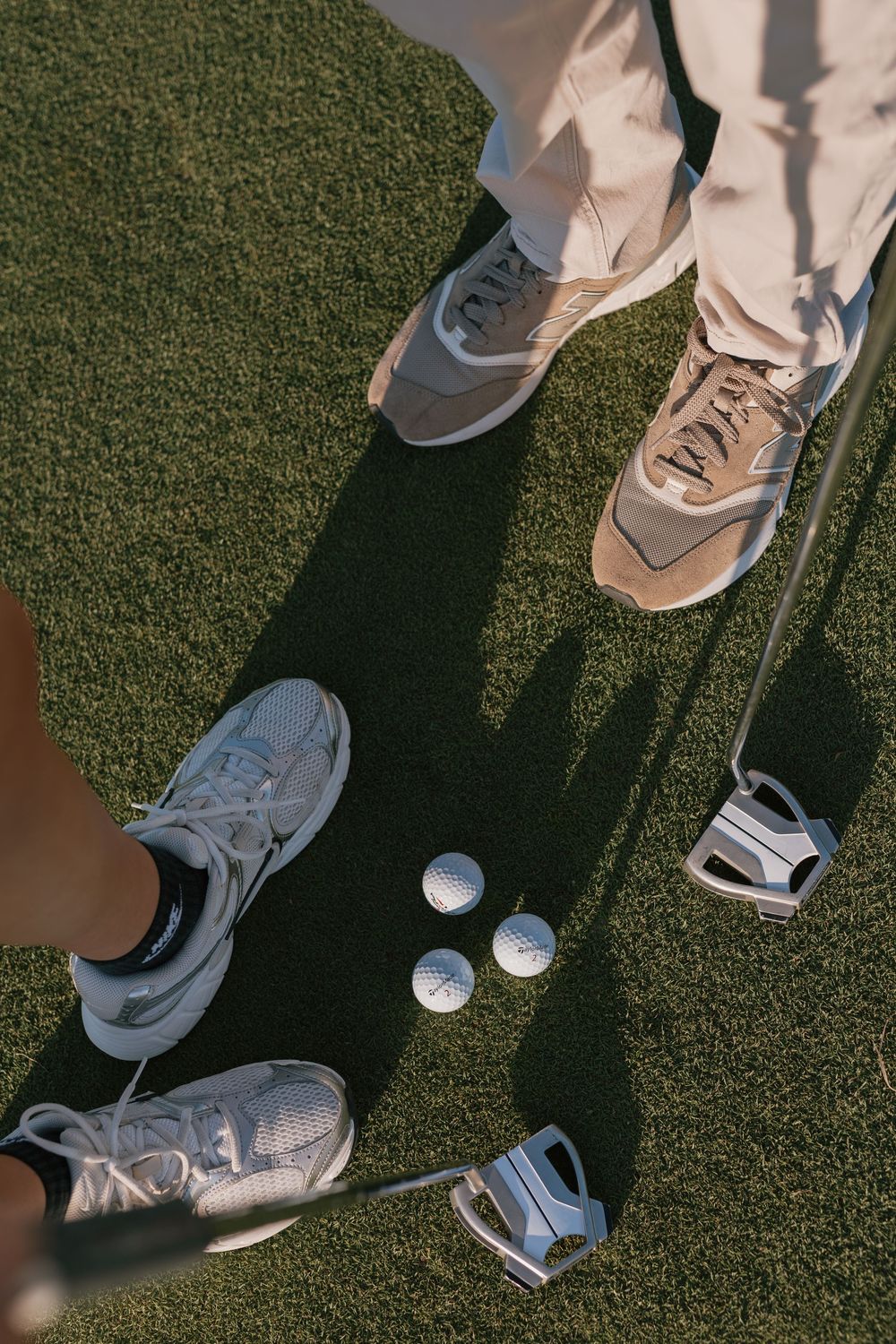 Two golfers standing on green grass with golf clubs and three white golf balls between their sneakers.