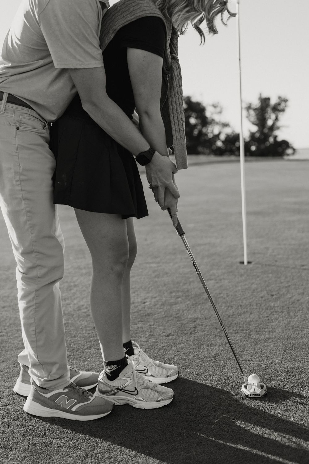 Black and white photo of a couple practicing golf putting together on a green, man guiding woman's stance.