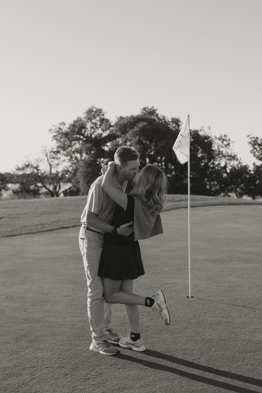 Black and white photo of a couple kissing on a golf course green beside a flag, captured during golden hour.