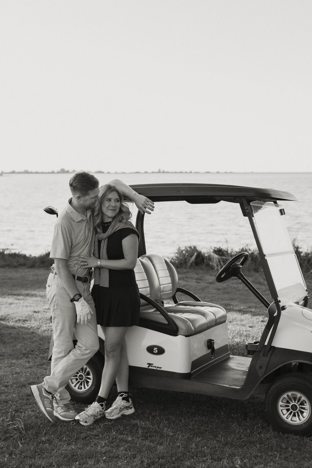 Black and white photo of a couple leaning against a golf cart near the water on a golf course.