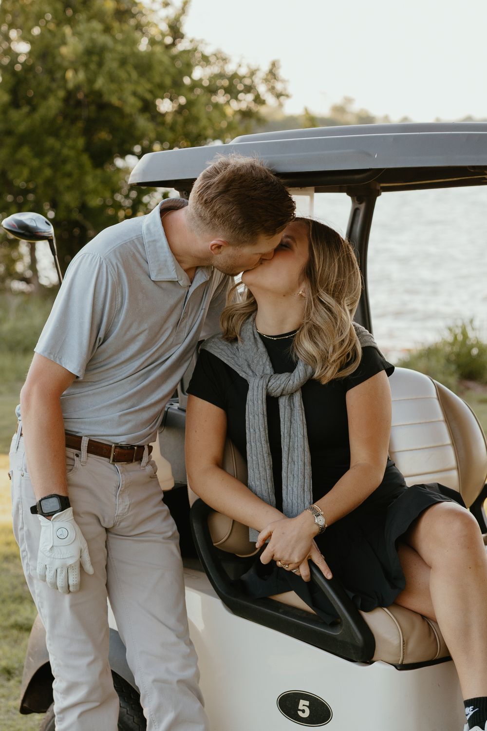Couple sharing a romantic kiss beside a golf cart near a serene lakeside, man wearing a golf glove and gray polo shirt.