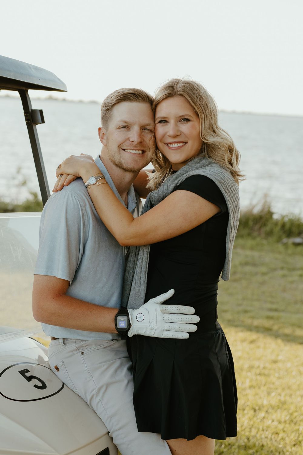 Happy couple embracing near a golf cart by the water, woman wearing a golf glove and gray scarf.
